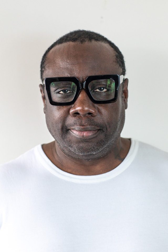 A person with a neutral expression, wearing large black glasses and a white shirt, stands against a plain background. professiona graphic design, motion design.