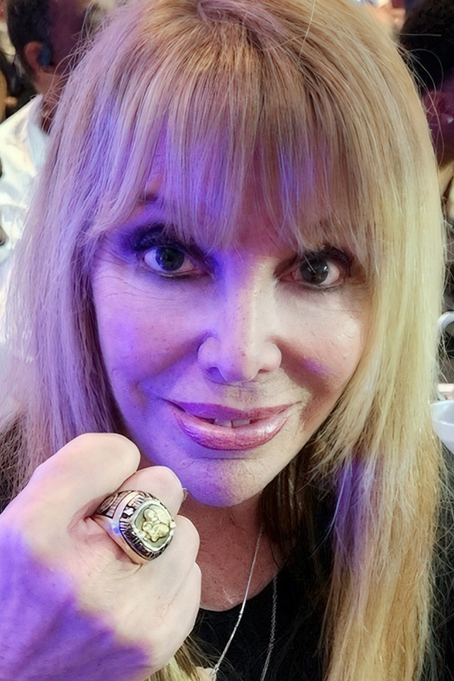 A woman with long blonde hair beams at the camera, raising her hand to showcase a ring that sparkles like a champions trophy. Bathed in purple-tinted light, she stands amidst a blurred, bustling crowd at the fever-pitched Tournament of Champions event.