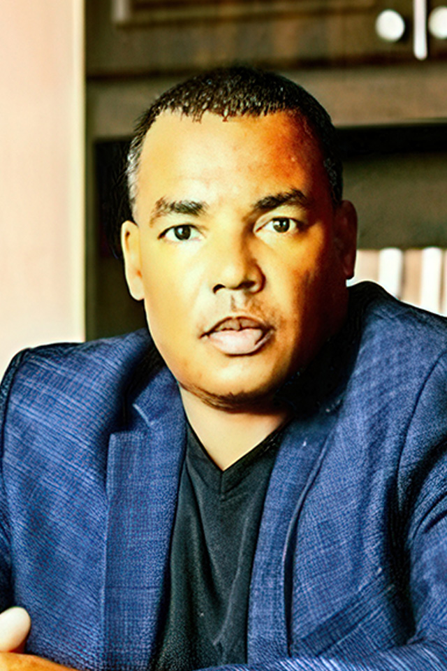 A man in a blue blazer and black shirt sits indoors with a bookshelf behind him, thoughtfully observing highlights from the tournament of champions.