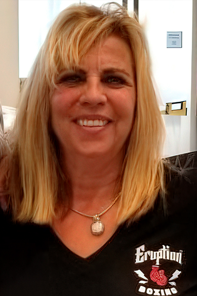 A woman with shoulder-length blonde hair smiles at the camera. She is wearing a black shirt with Eruption Boxing printed on it, along with a circular pendant necklace. The background shows a door and a white wall.