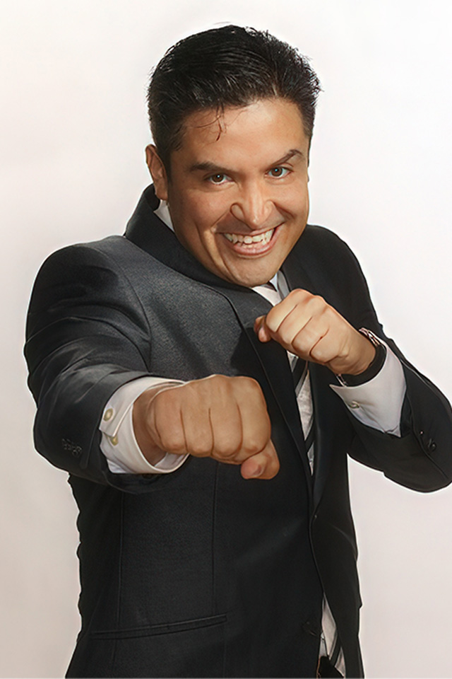 A man in a dark suit and white shirt playfully poses with his fists up, as if boxing, on a plain white background. With short dark hair and a broad smile, he exudes enthusiastic energy reminiscent of the lively vibe seen on esesports.tv.