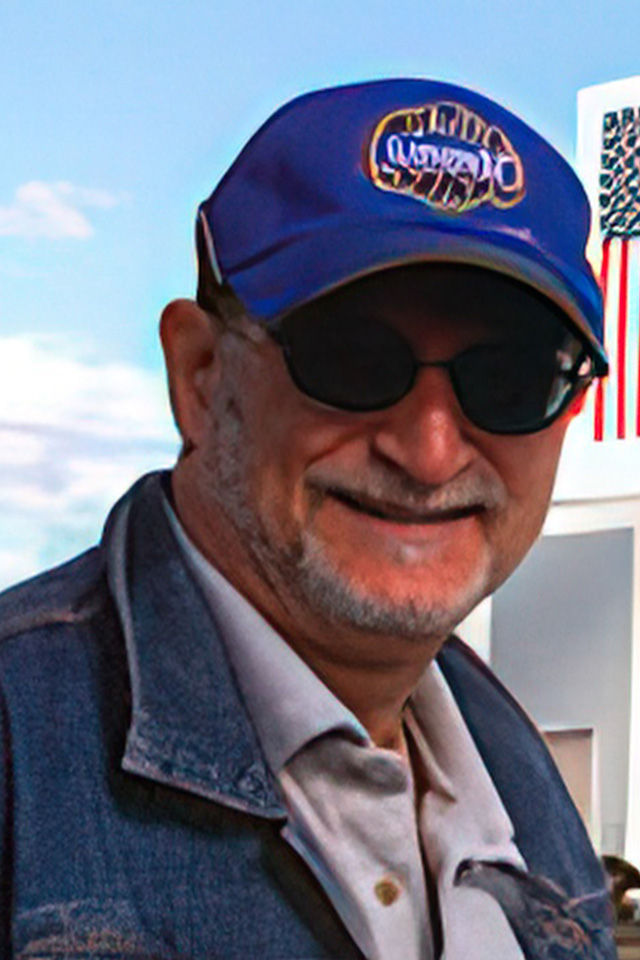 A man sporting a blue cap, dark sunglasses, and a denim jacket stands smiling under the partly cloudy sky in Mexico, with a section of a building visible behind him.