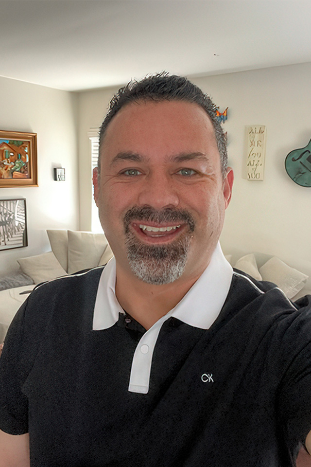 A man with short dark hair and a goatee smiles at the camera. Hes wearing a black polo shirt with a white collar, reminiscent of an announcer on esesports.tv. The background reveals a living room adorned with wall art, a guitar, and a couch with cushions.