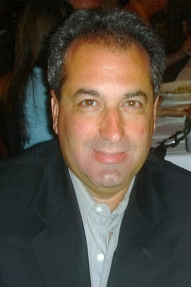 A man in a dark suit and light shirt smiles slightly at the camera, reminiscent of a champion after a boxing tournament. He has short, graying hair and is seated indoors, perhaps in Mexico or China, with blurred figures and objects in the background.