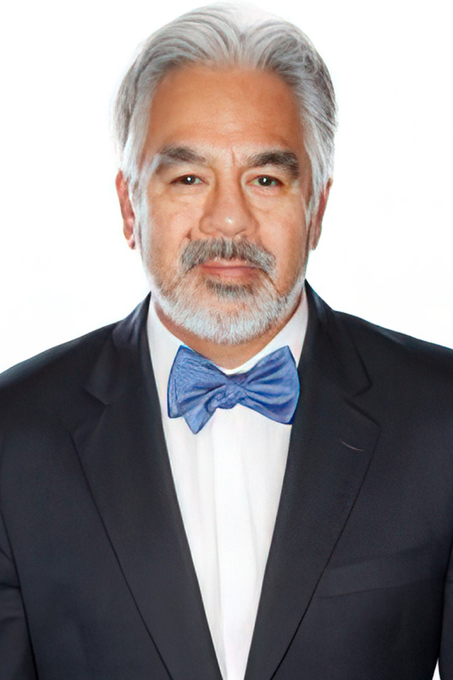 A man with gray hair and a beard is wearing a black suit, white shirt, and blue bow tie. He stands against a plain, light background, channeling the poised elegance of esesports.tv as he looks confidently at the camera.