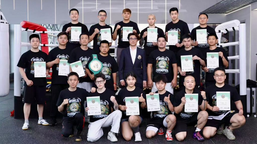 A group of 20 people, mostly in black T-shirts, pose in a gym holding certificates. One person holds a championship belt. Most are standing, with a few kneeling in the front row. Gym equipment is visible behind them.