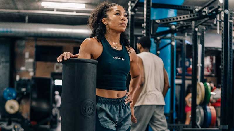 A woman in athletic wear stands in a gym, resting her hand on a foam roller and looking upward. Gym equipment and another person are visible in the background.