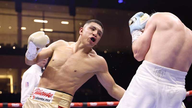 A male boxer in gold shorts throws a punch at his opponent, who is recoiling and covering his face while wearing white shorts and blue gloves, inside a boxing ring during a match.