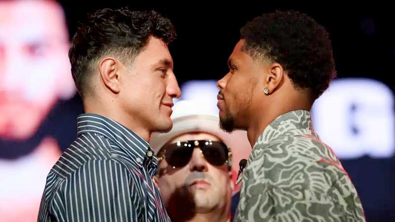 Two men stand face-to-face, staring each other down intensely, while a man in sunglasses and a white hat stands behind them. The background is dark with blurred text and images.
