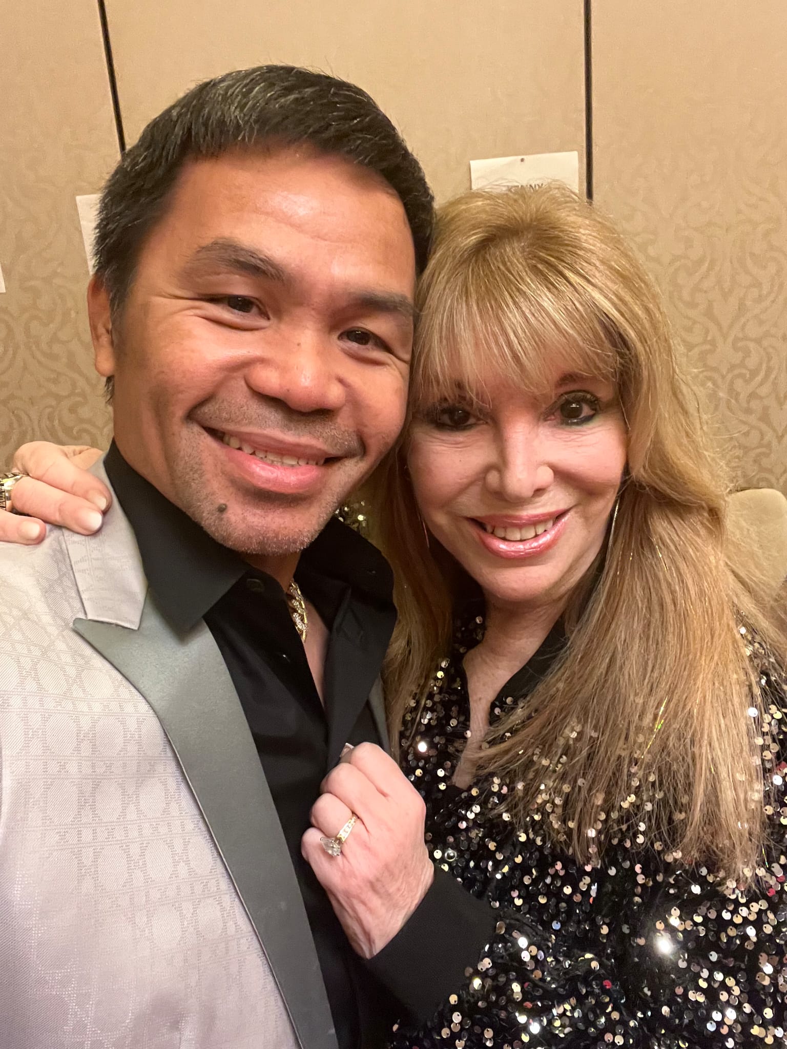 A man in a light suit and a woman in a sparkly black jacket smile together for a selfie, standing close in front of a tan, patterned wall.