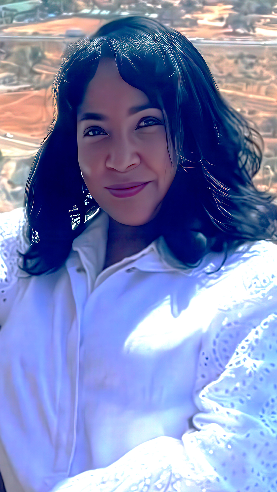 A woman with wavy dark hair smiles at the camera. She is wearing a white, textured blouse and is outdoors with blurred trees and a sunlit landscape in the background.