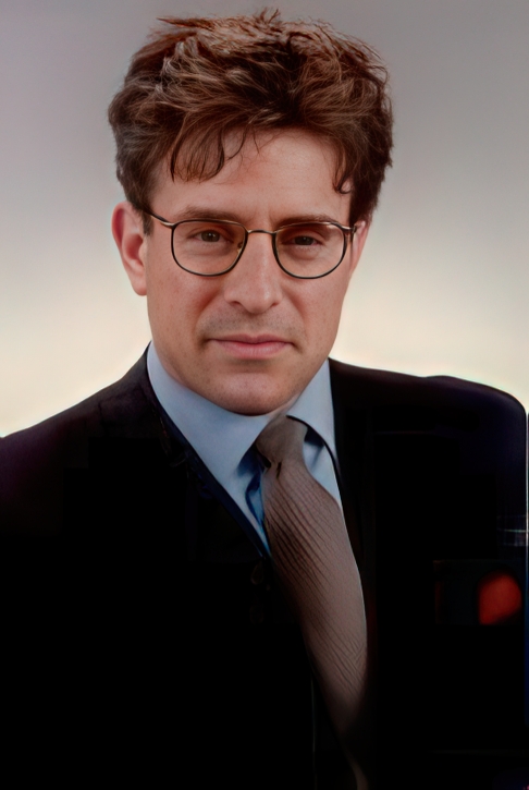 A man with short, wavy brown hair and glasses wears a dark suit, light shirt, and patterned tie, looking directly at the camera with a neutral expression against a light background.